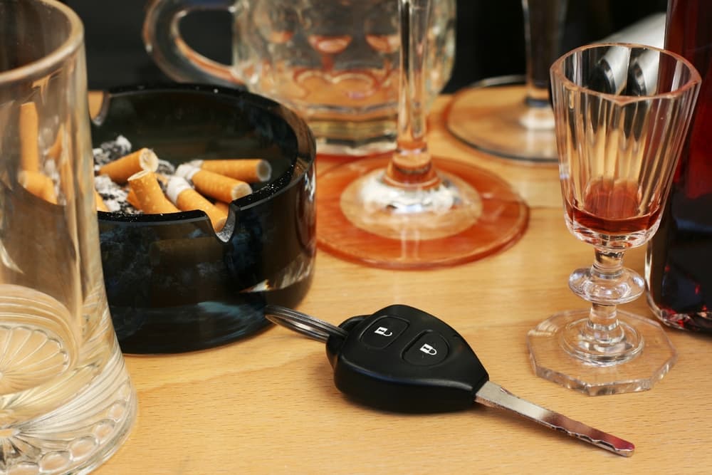 Car keys on the table full of empty glasses, bottles, full ashtray and spilled drink.