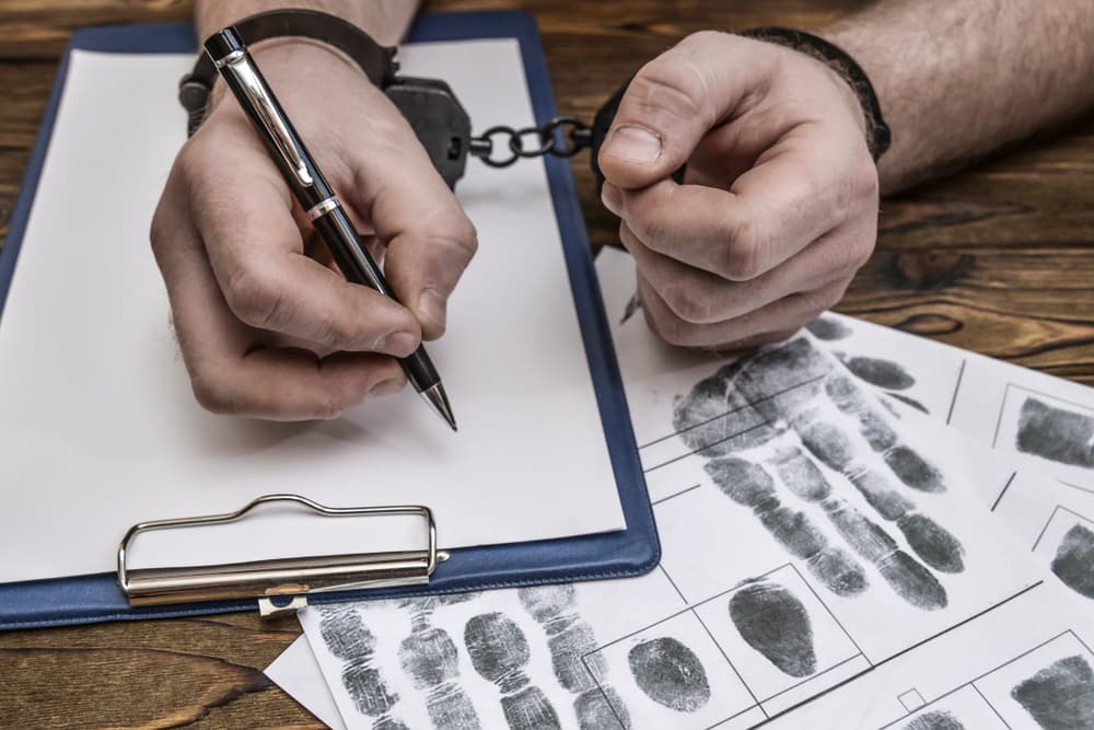 Handcuffed hands writing on clipboard, with fingerprint records spread on wooden table.