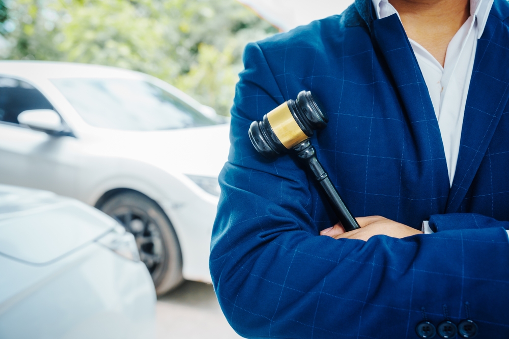A businessman in a suit and a lawyer holding a wooden gavel stand in front of a car, engaged in a discussion about legal issues such as citations, liability, and negligence related to a traffic violation.